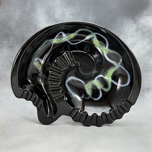 May include: A glossy black ceramic ashtray shaped like a coiled ammonite fossil. The surface features a decorative swirling pattern in shades of light green and pale blue, with integrated notches along the outer edge for resting items.