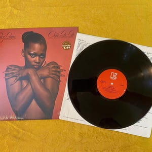 May include: Album cover for Suzi Lane's "Ooh La La". The cover features a woman with crossed arms against a red background. A vinyl record is next to the album cover, with a red label and white text. The album is from the Record Bar.