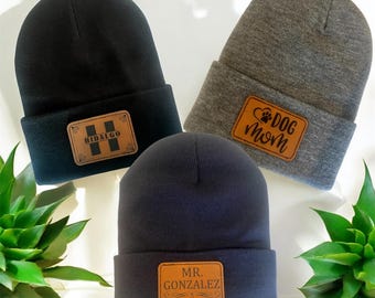 May include: Three knit beanies with leather patches. The black beanie has a patch that says "Hidalgo" with a stylized "H". The gray beanie has a patch that says "Dog Mom" with a paw print. The navy beanie has a patch that says "Mr. Gonzalez".