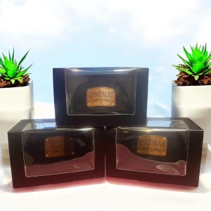 May include: Three black boxes with clear lids, each containing a black baseball cap with a gold-colored nameplate. The nameplates read "GONZALEZ", "SAMANTHA GARCIA", and "ESCOBAR".