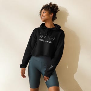 May include: Black cropped hoodie with a drawstring hood and long sleeves. The front features a white line drawing of breasts and the text "#NoSunBabes" in a handwritten font. The sleeve has a signature in white.