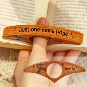 Könnte beinhalten: Hölzerner Buchseitenhalter mit dem Text "Just one more page" und einer Katzenillustration. Der Halter ist für einen Finger konzipiert und trägt den Namen "Sophia" sowie Stern- und Mondmotive. Ein zweiter Halter ist ebenfalls abgebildet.