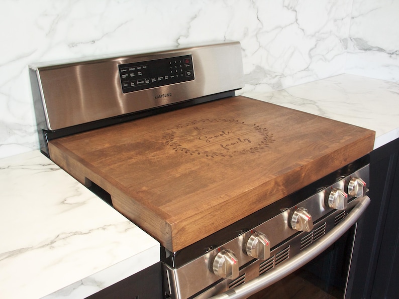 May include: A wooden stovetop cover with the text "Our Family" engraved in a wreath design. The cover is placed on a stainless steel stovetop.