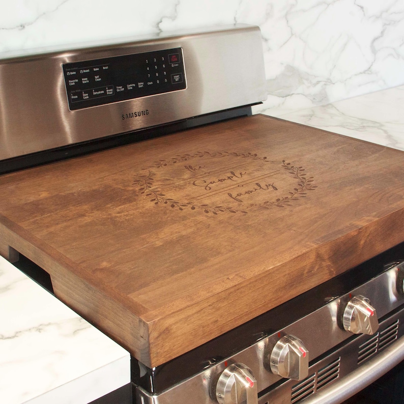 May include: A brown wooden stovetop cover with the text "The Sample Family" engraved in a wreath design.