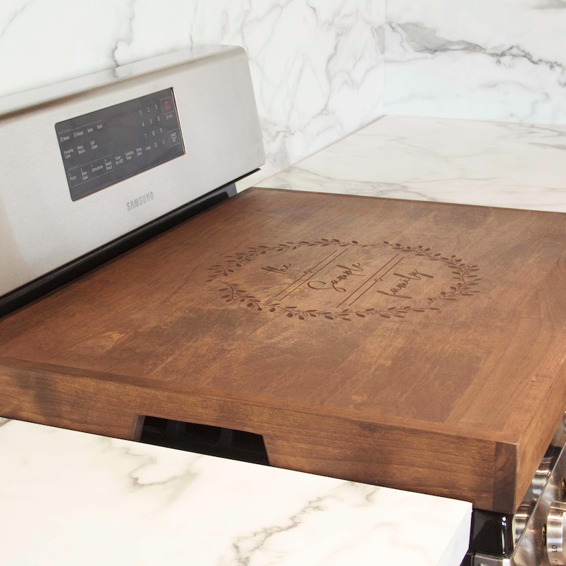 May include: A personalized wooden stovetop cover with the text "The [Last Name] Family" engraved in a wreath design.