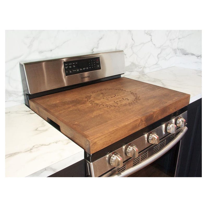 May include: A wooden stovetop cover with a decorative engraved wreath and the text "Gather family". The cover is brown and sits on top of a stainless steel stove.