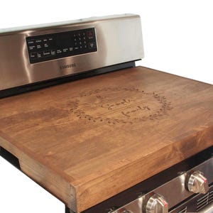 May include: A brown wooden stovetop cover with the text "The Sample Family" engraved in a wreath design.