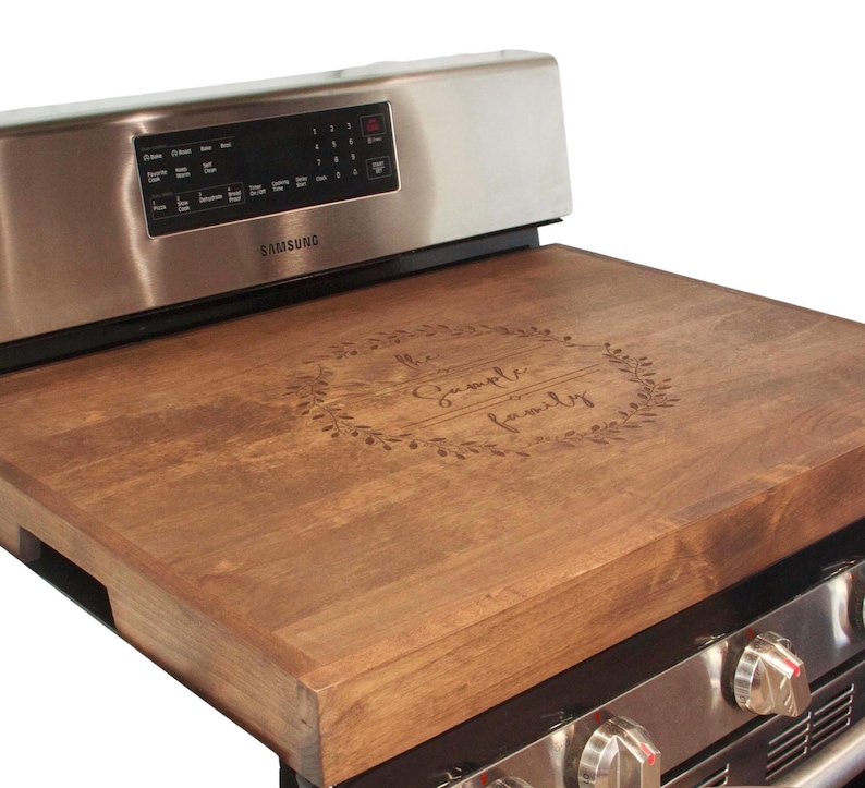 May include: A brown wooden stovetop cover with the text "The Sample Family" engraved in a wreath design.