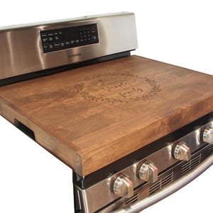 May include: A wooden stovetop cover with the text "Our Family" engraved in a wreath design. The cover is brown and sits on top of a stainless steel stove.