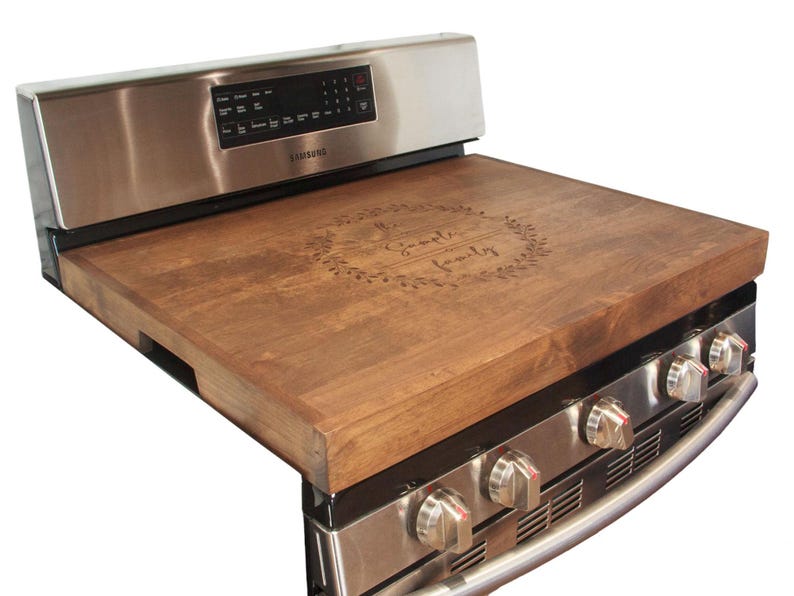 May include: A wooden stovetop cover with the text "Our Family" engraved in a wreath design. The cover is brown and sits on top of a stainless steel stove.