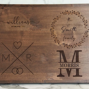 May include: A brown wooden cutting board with engraved text. The text reads "Williams Established 2022", "The Smith Family", and "M Morris". The board also features a heart, two rings, and a wreath.