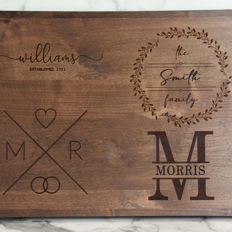 May include: A brown wooden cutting board with engraved text. The text reads "Williams Established 2022", "The Smith Family", and "M Morris". The board also features a heart, two rings, and a wreath.