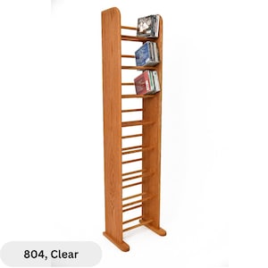 May include: Tall, wooden CD rack in a light brown finish. The rack has multiple horizontal shelves to hold CDs. The rack is holding several CDs. The text "804, Clear" is at the bottom of the image.