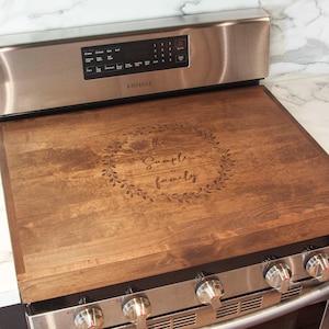 May include: A brown wooden stovetop cover with a wreath design and the text "The Sample Family" engraved on it. The cover is placed on top of a stainless steel stove.