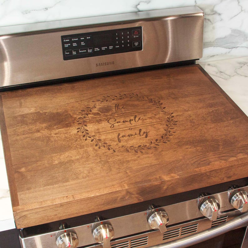 May include: A brown wooden stovetop cover with a wreath design and the text "The Sample Family" engraved on it. The cover is placed on top of a stainless steel stove.