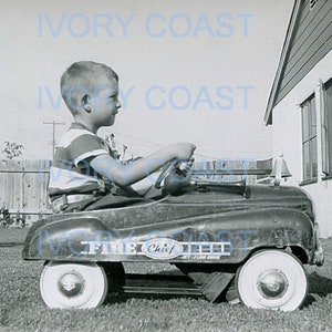 May include: A young boy in a striped shirt drives a red toy fire truck with a white steering wheel and white wheels. The toy truck has the words "Fire Chief" printed on the side.