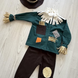 Pode incluir: Um conjunto de fantasia de espantalho composto por uma camisa de manga comprida verde-escura com franjas tipo palha nos punhos e gola, calças castanhas com franjas nos tornozelos e um chapéu pontiagudo castanho-escuro com remendos de tecido.