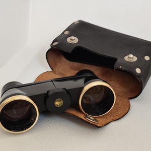 Könnte beinhalten: Schwarze Operngläser mit goldfarbenen Rändern und einem Fokus-Einstellknopf. Sie befinden sich in einem offenen schwarzen Lederetui mit braunem Innenfutter.