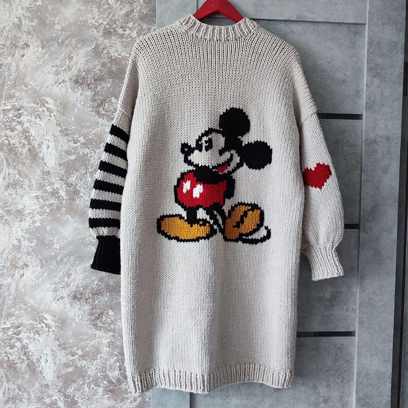 May include: A beige knitted cardigan with a Mickey Mouse design on the back. The cardigan has black and white striped sleeves with a red heart on one sleeve.