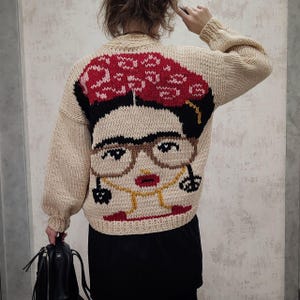 Pode incluir: Um su&eacute;ter de malha bege com uma imagem de malha colorida de Frida Kahlo nas costas. A imagem inclui seu monobrow ic&ocirc;nico, &oacute;culos e uma coroa de flores vermelhas.