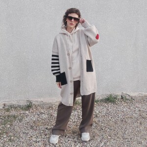 May include: A long-sleeved, beige cardigan with black and white striped sleeves and a black patch pocket. The cardigan has a red heart patch on the right sleeve and is worn with brown pants.