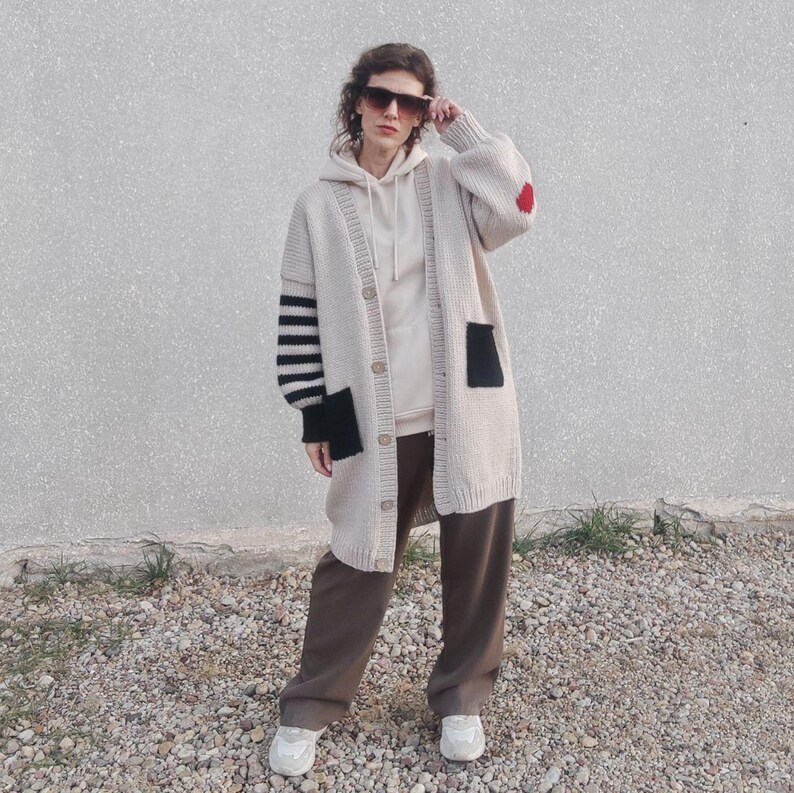 May include: A long-sleeved, beige cardigan with black and white striped sleeves and a black patch pocket. The cardigan has a red heart patch on the right sleeve and is worn with brown pants.