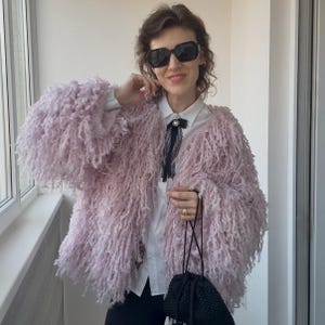 Puede incluir: Chaqueta peluda de color lila claro con mangas largas. La chaqueta se lleva sobre una camisa blanca con cuello y un lazo negro. La persona lleva gafas de sol negras y sostiene una bolsa negra con cordón.