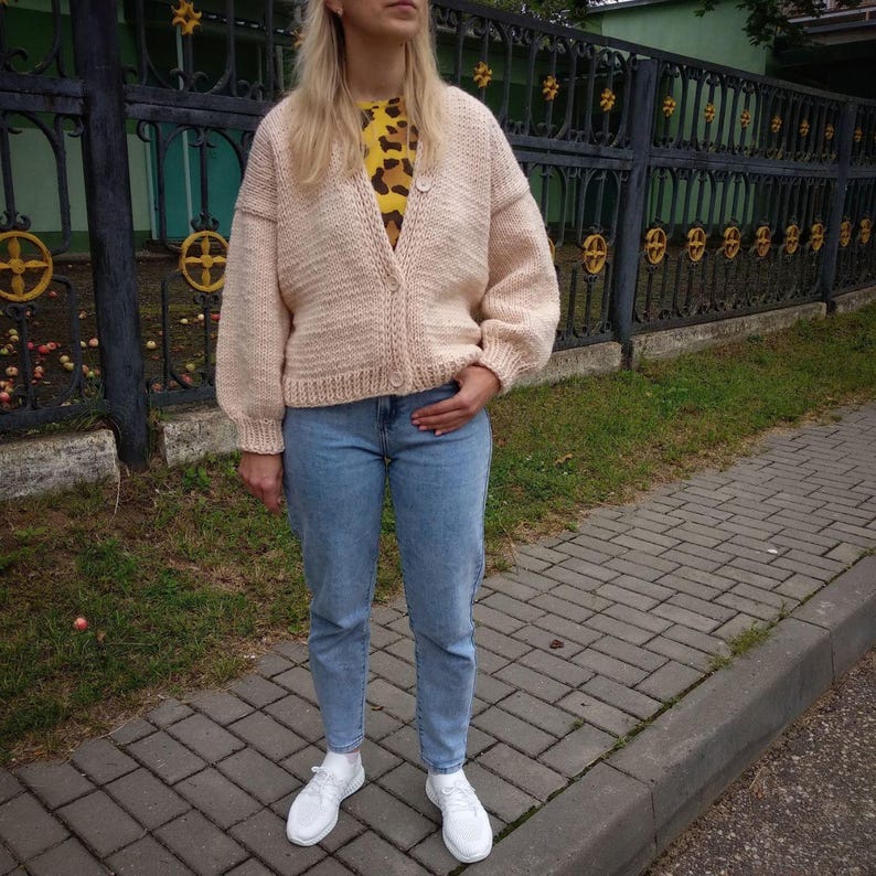 Pode incluir: Um cardigan de malha rosa claro com fecho de bot&otilde;es. O cardigan tem mangas compridas e um caimento solto. A modelo est&aacute; usando o cardigan sobre uma camisa estampada e cal&ccedil;as de ganga azuis.