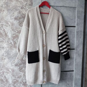 May include: A long, beige cardigan with black patch pockets and striped sleeves. The cardigan has a button closure and a V-neck.