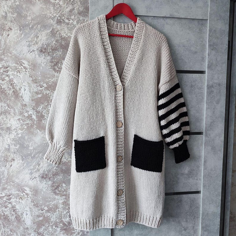 May include: A long, beige cardigan with black patch pockets and striped sleeves. The cardigan has a button closure and a V-neck.