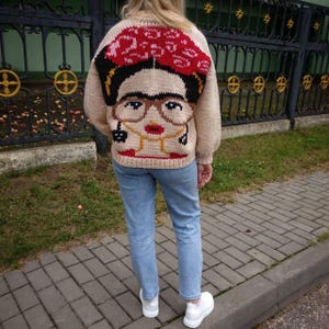 Pode incluir: Um su&eacute;ter de tric&ocirc; bege com um retrato colorido de Frida Kahlo na frente. O su&eacute;ter apresenta uma faixa de cabe&ccedil;a vermelha, cabelo preto, &oacute;culos e um colar de ouro.