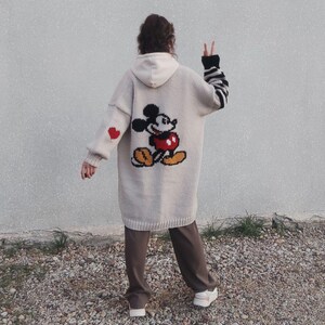 May include: A long, beige cardigan with a Mickey Mouse graphic on the back. The cardigan has red hearts on the sleeves and a black and white striped pattern on one sleeve.