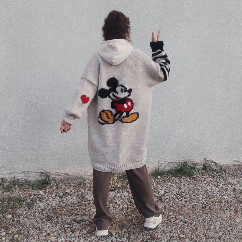 May include: A long, beige cardigan with a Mickey Mouse graphic on the back. The cardigan has red hearts on the sleeves and a black and white striped pattern on one sleeve.