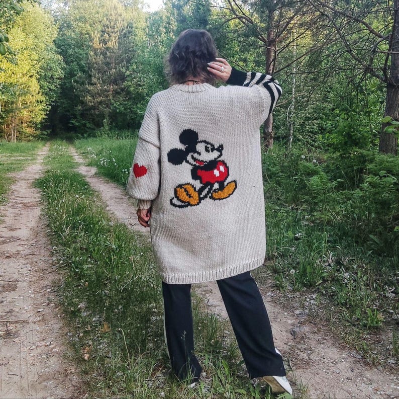 May include: A white knitted cardigan with a Mickey Mouse design on the back. The cardigan has red hearts on the sleeves and a black and white striped trim on the right sleeve.