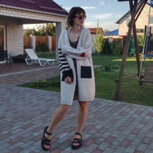 May include: A long-sleeved, light gray cardigan with black and white striped sleeves. The cardigan has a red heart on the front and a black patch pocket. The model is wearing black sandals.