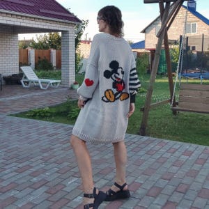 May include: A light gray knitted cardigan with a Mickey Mouse graphic on the back. The cardigan has a black and white striped sleeve and a red heart on the other sleeve.