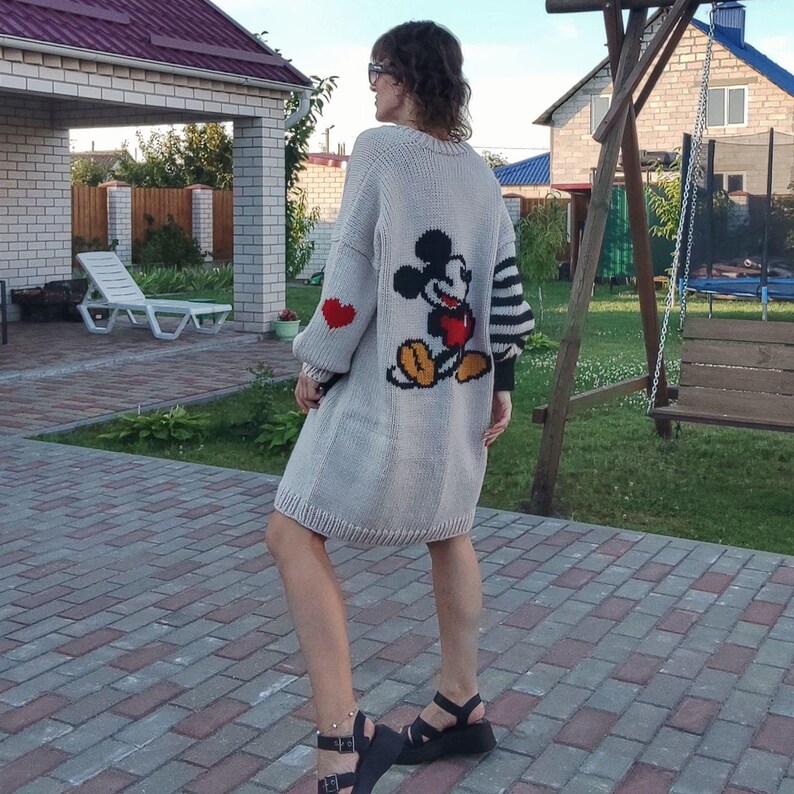 May include: A light gray knitted cardigan with a Mickey Mouse graphic on the back. The cardigan has a black and white striped sleeve and a red heart on the other sleeve.