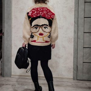 Pode incluir: Um su&eacute;ter de tric&ocirc; cor creme com um retrato colorido de Frida Kahlo nas costas. O su&eacute;ter tem gola redonda e mangas compridas.