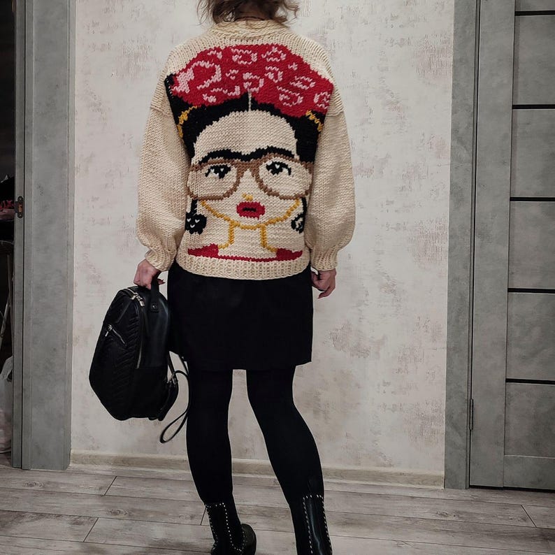 Pode incluir: Um su&eacute;ter de tric&ocirc; cor creme com um retrato colorido de Frida Kahlo nas costas. O su&eacute;ter tem gola redonda e mangas compridas.