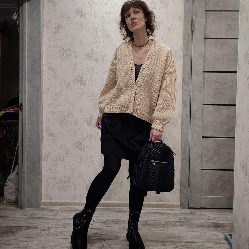 Pode incluir: Uma mulher vestindo um cardig&atilde; de tric&ocirc; bege com uma saia preta e meias pretas. Ela est&aacute; carregando uma mochila acolchoada preta.