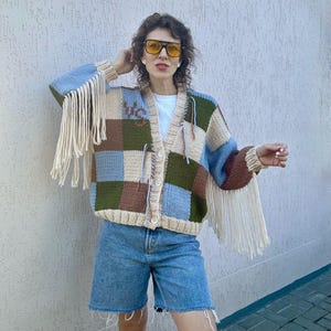 May include: A patchwork cardigan in shades of blue, brown, green, and beige with fringe sleeves. The cardigan has a V-neck and button closure. The sleeves are a solid brown color. The cardigan is worn with denim shorts.