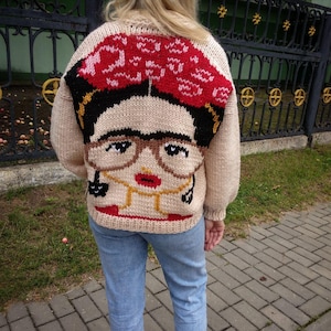 May include: A beige knitted jumper with a colourful knitted image of Frida Kahlo on the back. The image includes a black headband, a red flower, glasses, and a gold necklace.