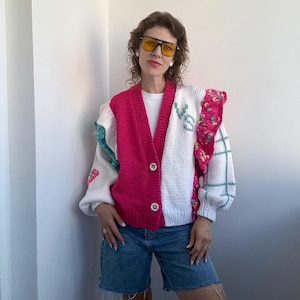 Op de afbeelding: Een colorblock vest in wit en felroze met bloemen en groene accenten. De trui heeft een V-hals, knoopsluiting en het woord "VE" geborduurd op de voorkant. De mouwen zijn lang en poofy en het kledingstuk wordt gedragen met een spijkerbroekshort.