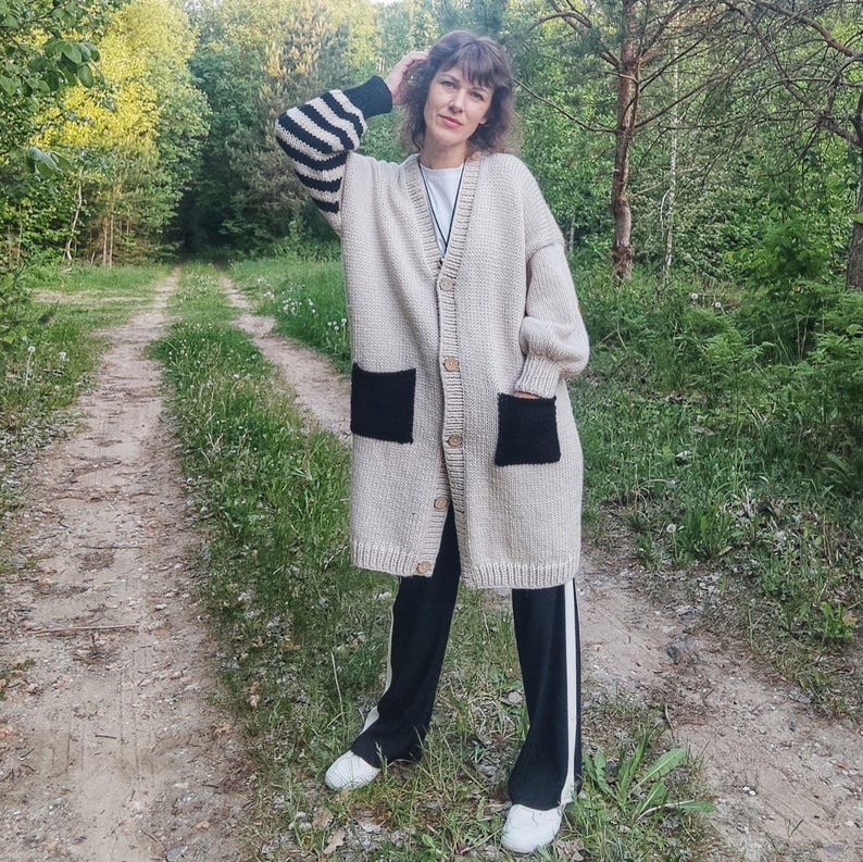 May include: A beige cardigan with black and white striped sleeves and black patch pockets. The cardigan is open and the person wearing it is standing on a path in a forest.