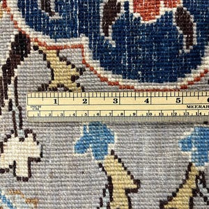 Puede incluir: Primer plano de una secci&oacute;n de una alfombra con un dise&ntilde;o floral en tonos azules, beige y marr&oacute;n. Una regla se coloca sobre la alfombra, mostrando medidas en pulgadas y cent&iacute;metros. La alfombra tiene un fondo gris.