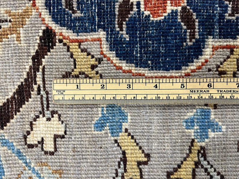 Puede incluir: Primer plano de una secci&oacute;n de una alfombra con un dise&ntilde;o floral en tonos azules, beige y marr&oacute;n. Una regla se coloca sobre la alfombra, mostrando medidas en pulgadas y cent&iacute;metros. La alfombra tiene un fondo gris.