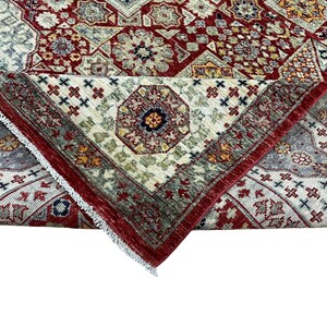 Puede incluir: Una alfombra plegada con un intrincado dise&ntilde;o geom&eacute;trico. La alfombra presenta una rica paleta de colores de rojo, crema y toques de verde y dorado. El dise&ntilde;o incluye motivos florales y geom&eacute;tricos repetidos, con un borde rojo.