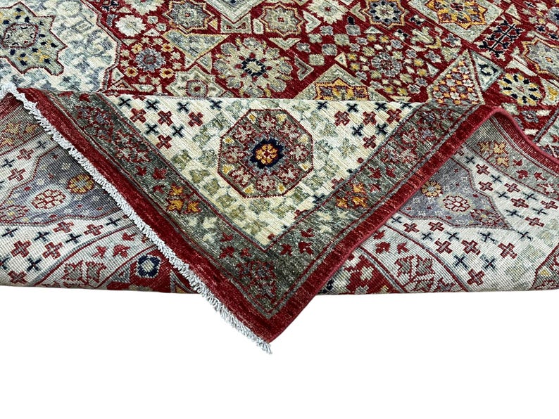 Puede incluir: Una alfombra plegada con un intrincado dise&ntilde;o geom&eacute;trico. La alfombra presenta una rica paleta de colores de rojo, crema y toques de verde y dorado. El dise&ntilde;o incluye motivos florales y geom&eacute;tricos repetidos, con un borde rojo.