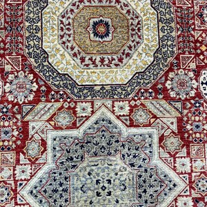 Puede incluir: Primer plano de una alfombra detallada y ornamentada con un dise&ntilde;o octogonal central. La alfombra presenta una rica paleta de colores rojo, beige, azul marino y dorado. Los intrincados patrones florales y geom&eacute;tricos crean una apariencia tradicional y texturizada.