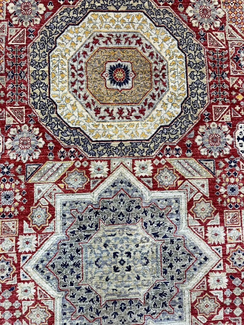 Puede incluir: Primer plano de una alfombra detallada y ornamentada con un dise&ntilde;o octogonal central. La alfombra presenta una rica paleta de colores rojo, beige, azul marino y dorado. Los intrincados patrones florales y geom&eacute;tricos crean una apariencia tradicional y texturizada.